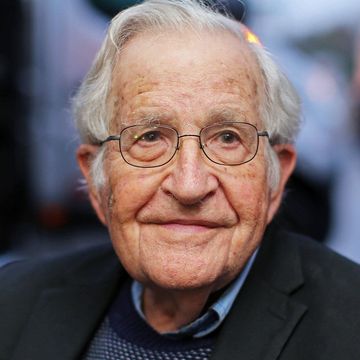 noam chomsky smiles and looks past the camera, he wears glasses, a black suit jacket, a blue collared shirt and a blue and white sweater