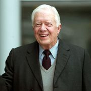jimmy carter smiles and waves to the camera, he is wearing a blue and white striped collared shirt with a navy and dark red diagonal striped tie, a light gray v neck sweater is on top of the shirt and tie, and he also wears a dark gray suit jacket