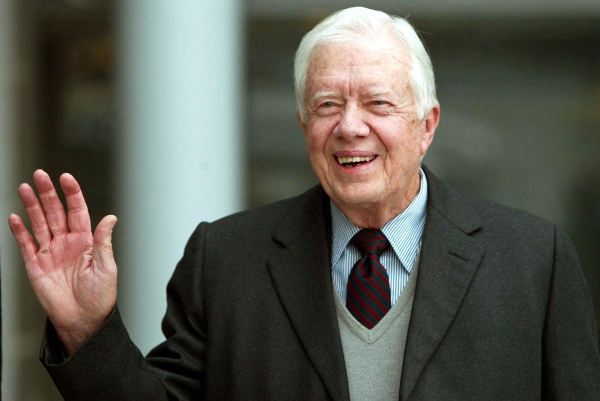 jimmy carter smiles and waves to the camera, he is wearing a blue and white striped collared shirt with a navy and dark red diagonal striped tie, a light gray v neck sweater is on top of the shirt and tie, and he also wears a dark gray suit jacket