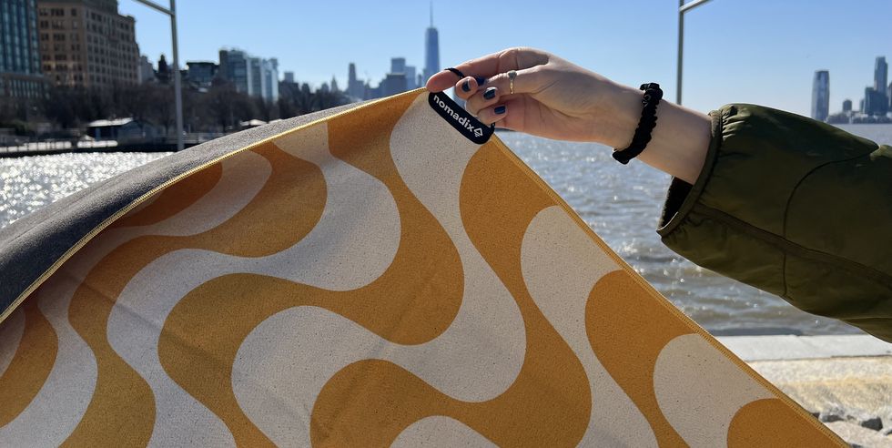 a yellow and white patterned nomadix original towel being held up in the air on the beach, good housekeeping's best beach towels