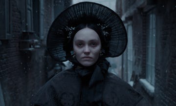 a figure in a dark ornate outfit and a large hat standing in a snowcovered alleyway