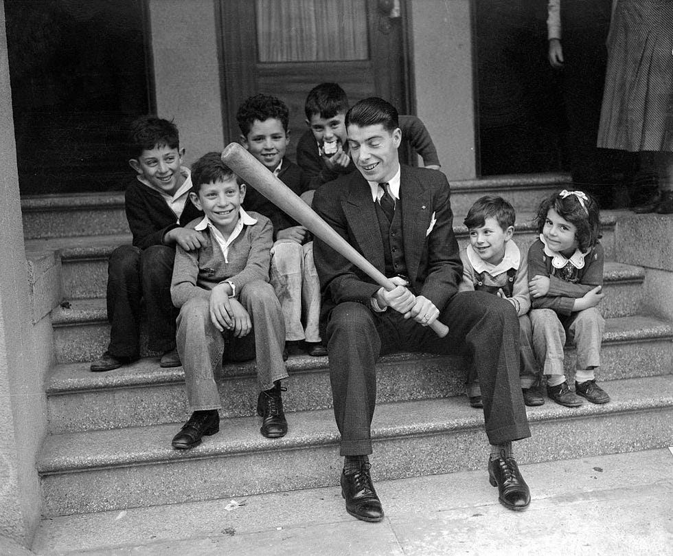 joe dimaggio sits with children on steps outside a home and holds a bat, everyone smiles