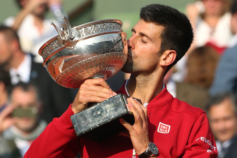 novak djokovic holds a silver trophy as he kisses it, he wears a red jacket and watch