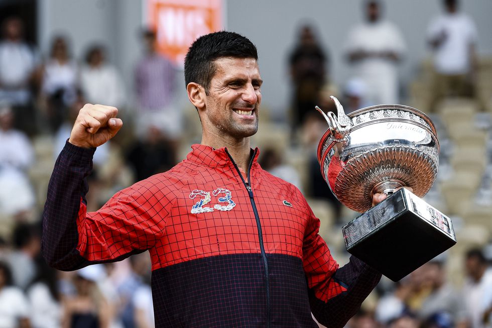 novak djokovic pumps his fist and holds the french open trophy, he wears a red and black jacket with the number 23 on the front