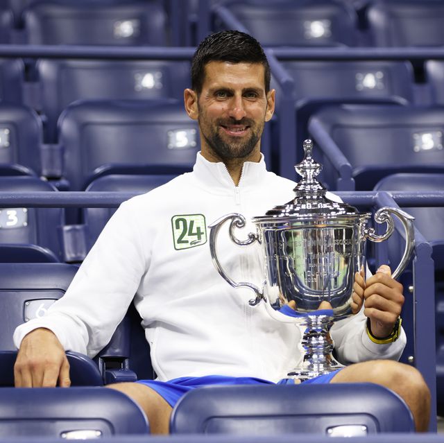 novak djokovic sits in a stadium seat and smiles at the camera, he holds a large silver trophy that rests on his leg and wears a white jacket with the number 24 on it and blue shorts
