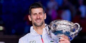 novak djokovic holds the australian open trophy and smiles at the camera, he wears a white jacket