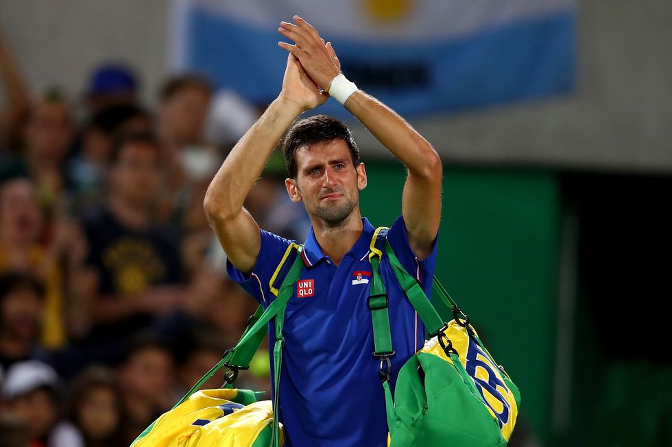 novak djokovic claps his has over his head, he carries two tennis bags and wears a blue polo shirt, his face is red and scrunched from crying