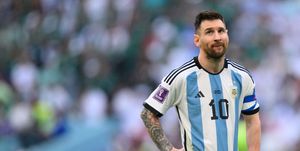 lionel messi stands with his hands on his hips during a game of the 2022 world cup, he is wearing a white and blue striped argentina national team uniform shirt