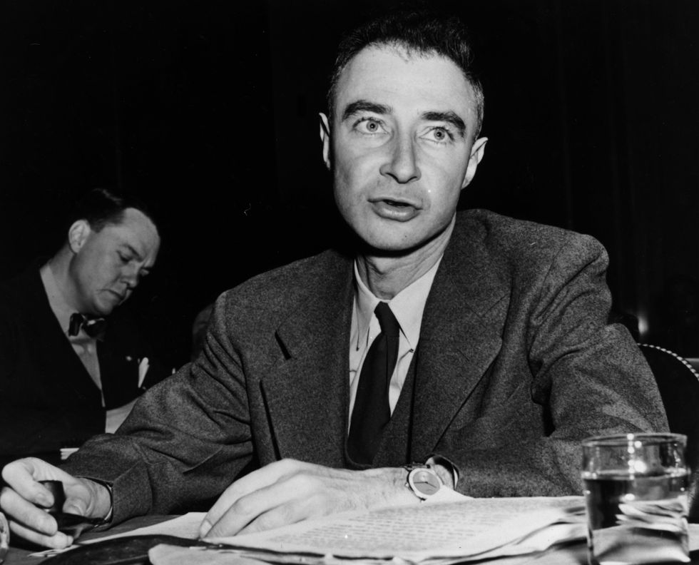 a black and white photo of j robert oppenheimer, wearing a suit and sitting at a table, speaking to someone off camera