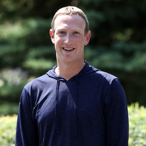 mark zuckerberg stands outside and smiles at the camera, he is wearing a long sleeve navy blue hoodie