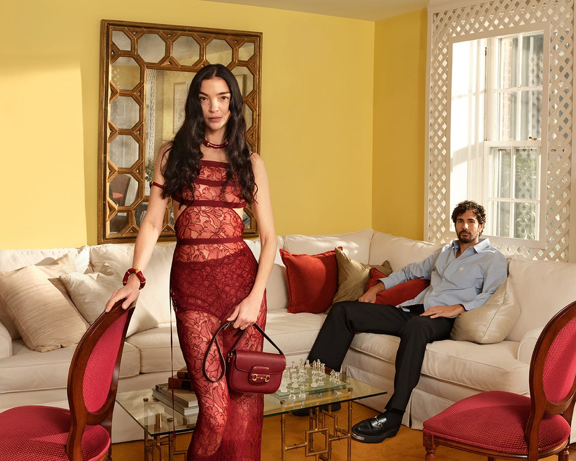 woman in a red lace dress stands in a living room with a man seated on a sofa