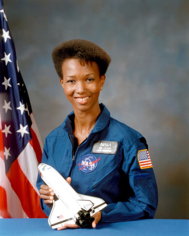 mae c jemison smiles and poses for a photo, she holds a minature space shuttle in front of her and wears a blue nasa uniform, an american flag stands in the background to the left