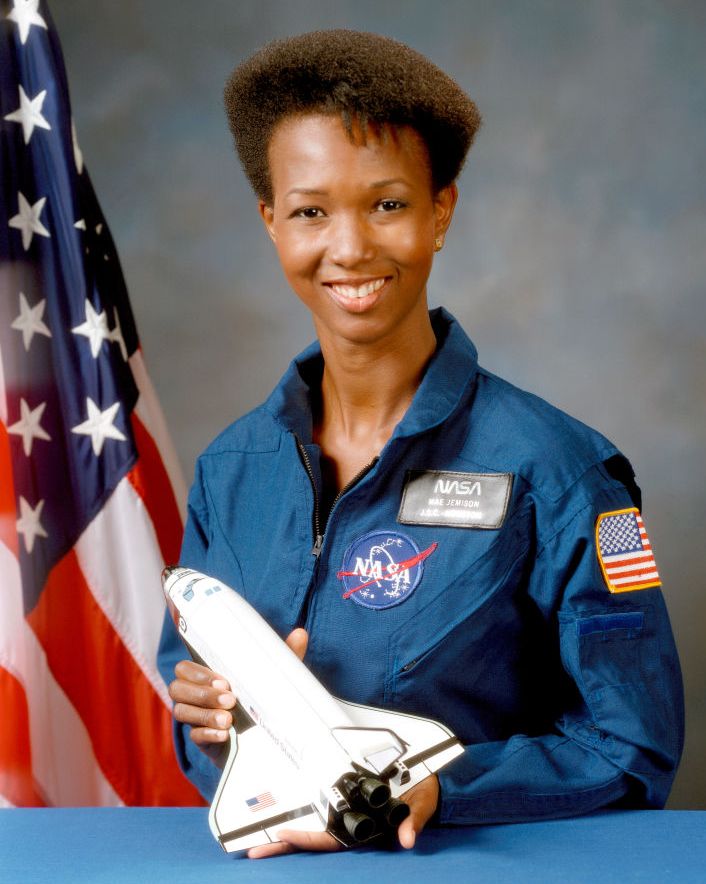 mae c jemison smiles and poses for a photo, she holds a minature space shuttle in front of her and wears a blue nasa uniform, an american flag stands in the background to the left