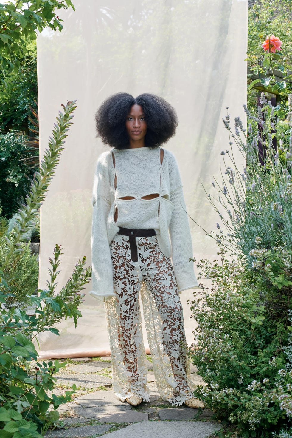 a person wearing a stylish outfit consisting of a sweater with cutouts and lace pants set in a lush garden