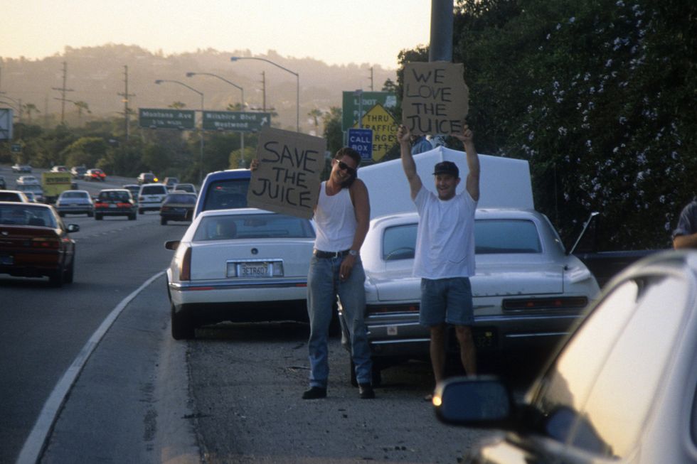 two men stand on a highway shoulder and hold cardboard signs with messages, cars drive past