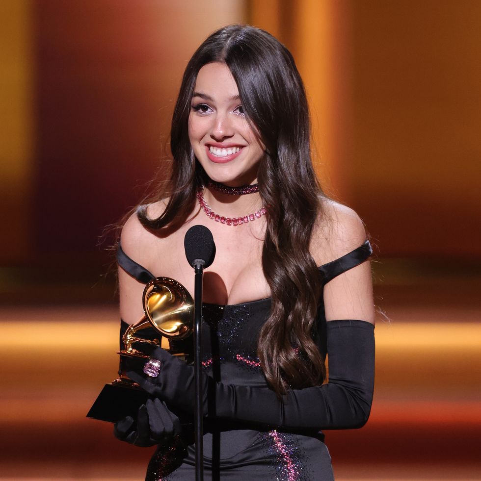 olivia rodrigo stands at the microphone and holds a grammy trophy, she smiles and wears a black dress with off the shoulder sleeves and long black gloves
