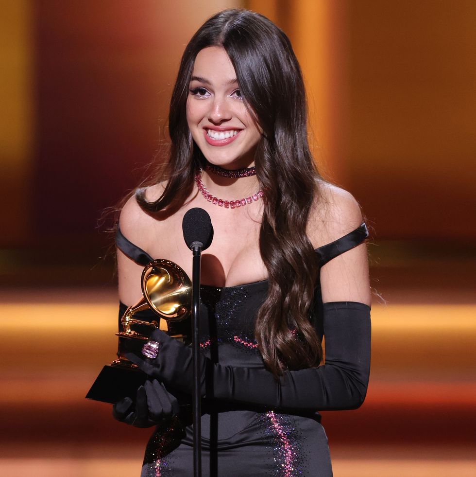olivia rodrigo stands at the microphone and holds a grammy trophy, she smiles and wears a black dress with off the shoulder sleeves and long black gloves