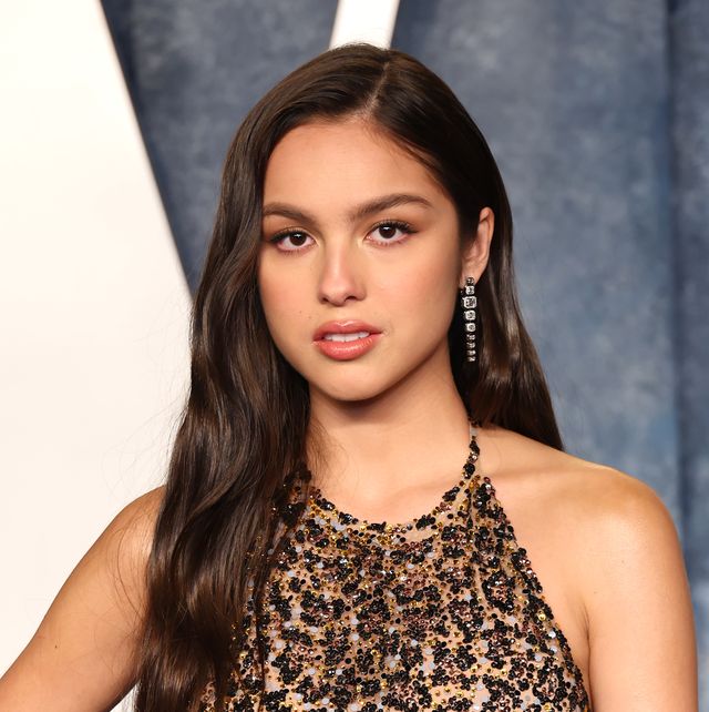 olivia rodrigo looks at the camera, she wears a cheetah inspired halter top dress with sequins and dangling earrings