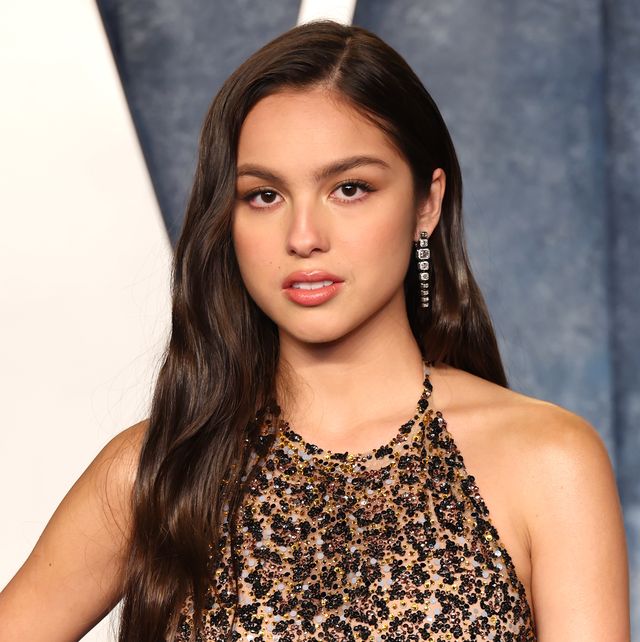 olivia rodrigo looks at the camera, she wears a cheetah inspired halter top dress with sequins and dangling earrings