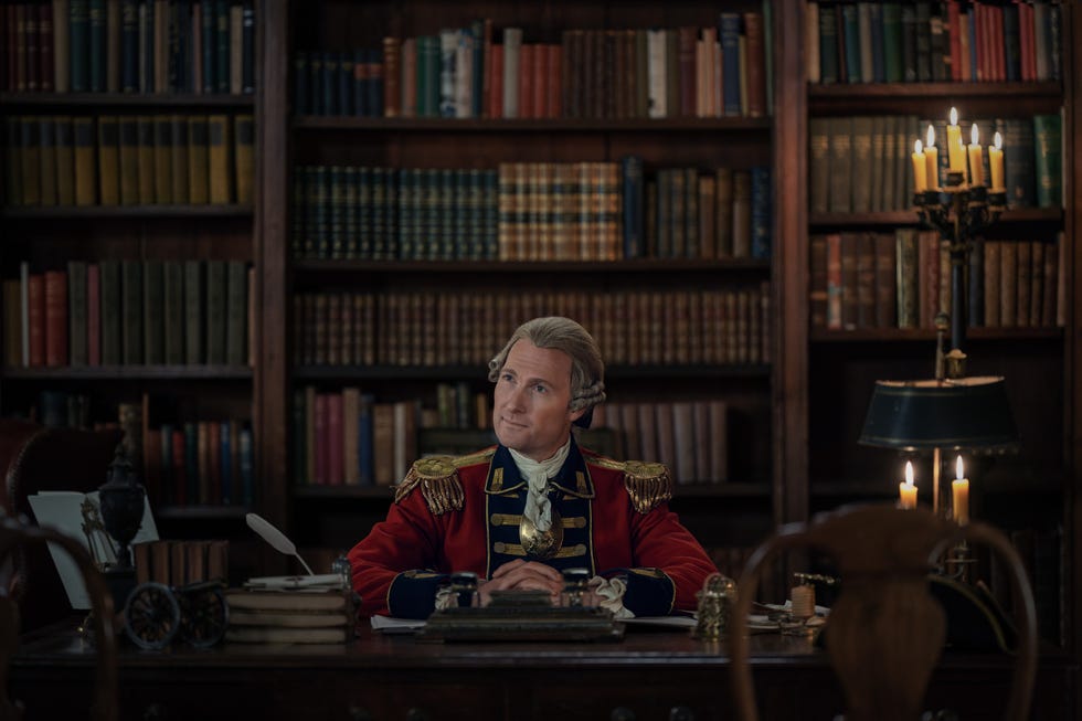 a historical figure in military attire seated at a desk surrounded by books in a library setting