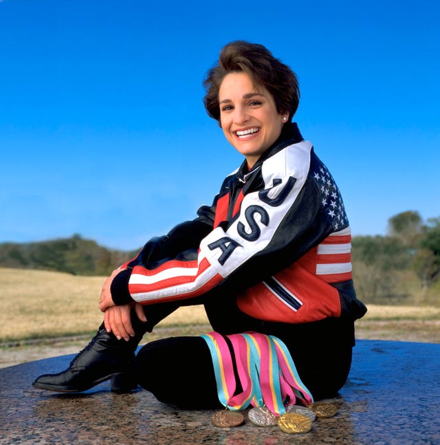 mary lou retton smiles and sits outside, she wears a usa jacket, black pants, and black boots, five medals and their multicolor lanyard rest on her thigh and the stone she sits on