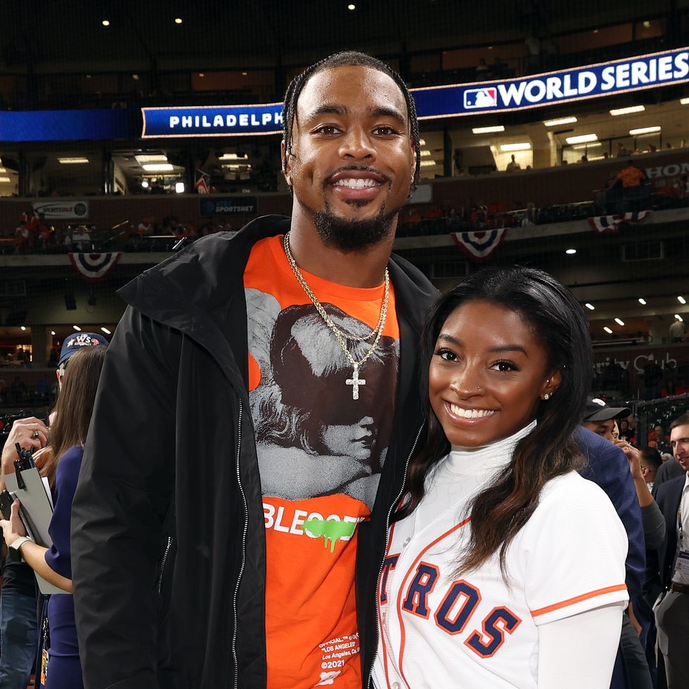 jonathan owens and simone biles hug and smile for a photo while standing inside a baseball stadium, he wears a black jacket over an orange t shirt, she wears a white houston astros baseball jersey over a white turtleneck
