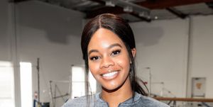 gabby douglas stands inside a gymnastics training facility and smiles at the camera with one hand on her hip, she wears a gray long sleeve top with her hair in a ponytail