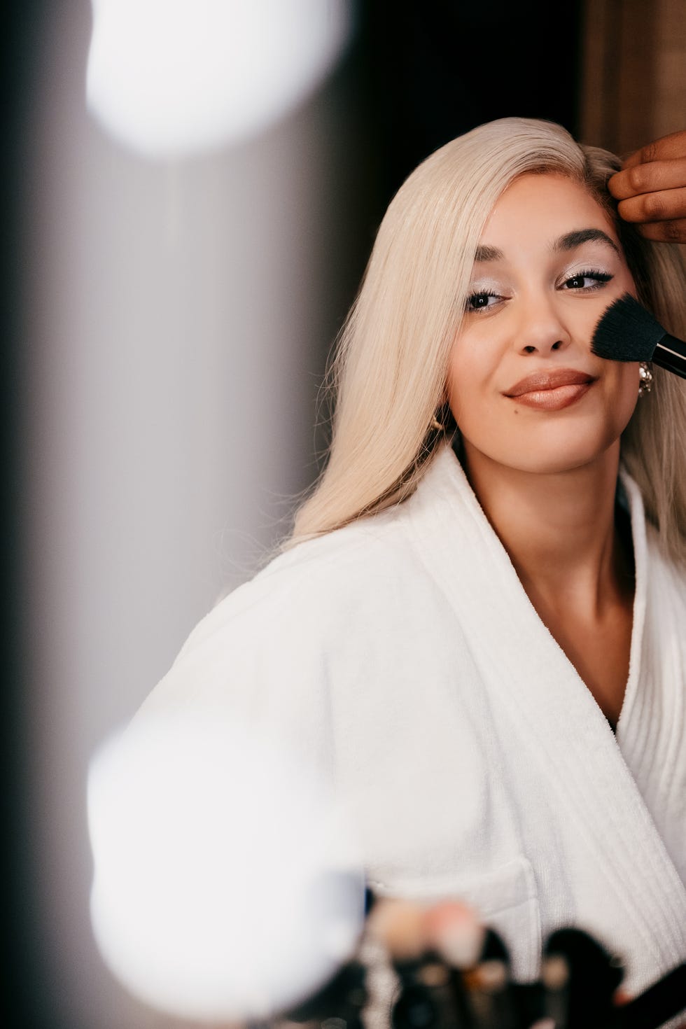 a person with long blonde hair getting makeup applied while wearing a white robe