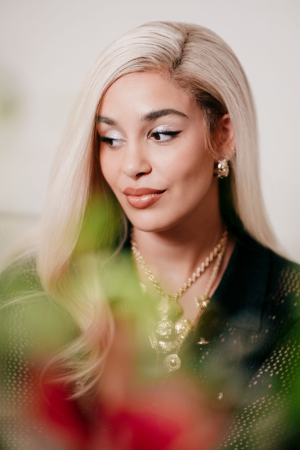 a person wearing multiple gold necklaces with long blonde hair partially obscured by green plants