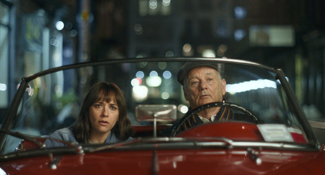 A convertible is parked at night, two passengers are sitting in the front seat, the driver is wearing a cap and a striped shirt, while the passenger has long hair and is dressed in a denim-like robe, blurred city lights illuminate the background, creating something of a gentle atmosphere around the scene