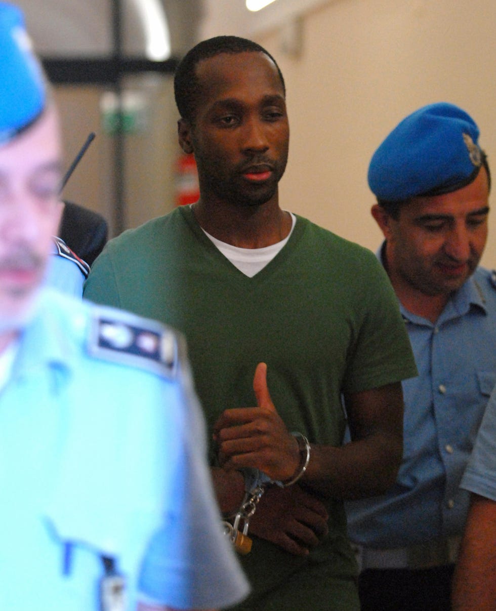 a man in handcuffs walks in a hallway surrounded by several police officers, he wears a green short sleeve sweater