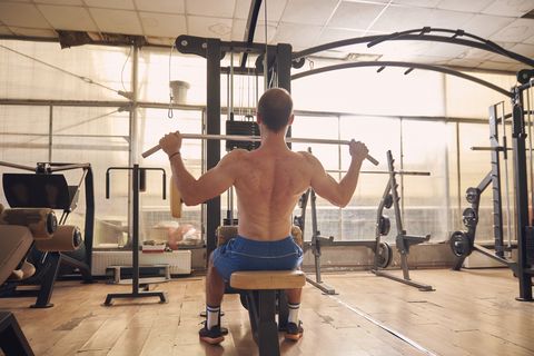 one young man, shirtless, back lat pulldown exercise, sitting multistationary machine  in old beaten up gym interior. full length shot, rear view.