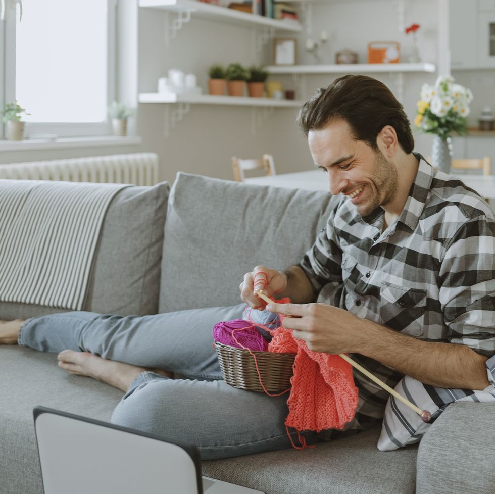 a man knitting on the couch in front of a computer screen