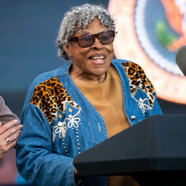 Opal Lee, the “Godmother of Juneteenth” opal lee, wearing a denim designed coat, yellow shirt, and sunglasses, speaks at a podium with two microphones and the us presidential seal, standing next to a laughing kamala harris