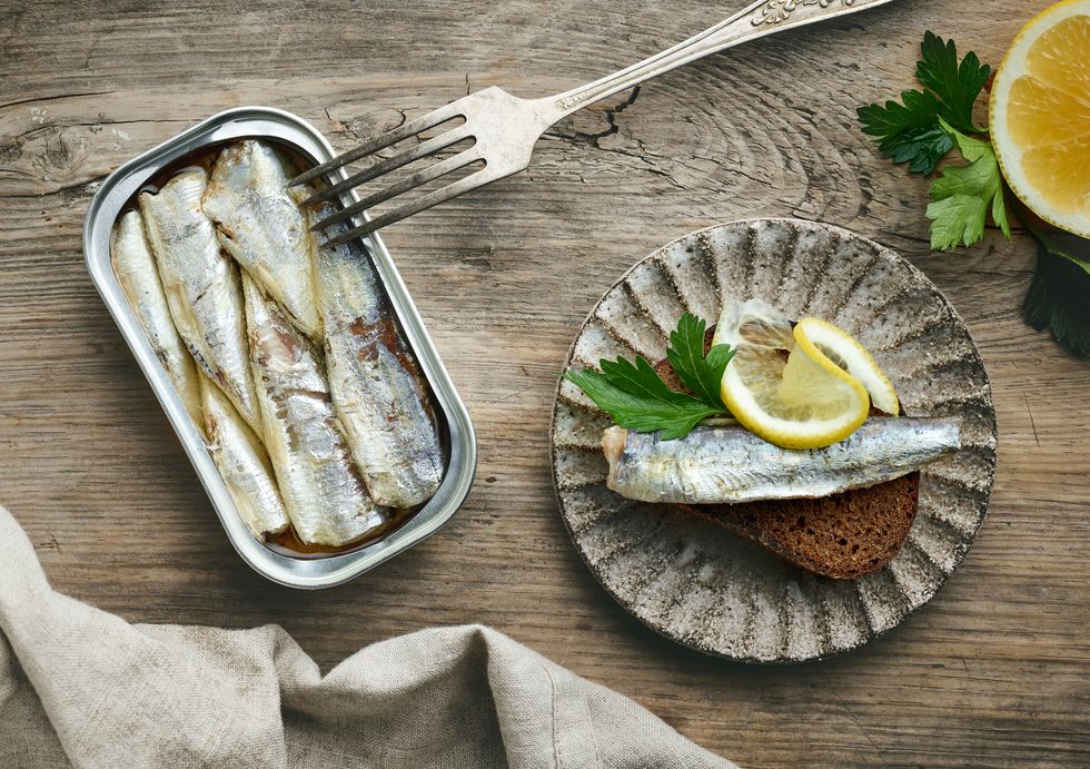 an open can of sardines on a table top next to a plate with sardine toast and lemon.