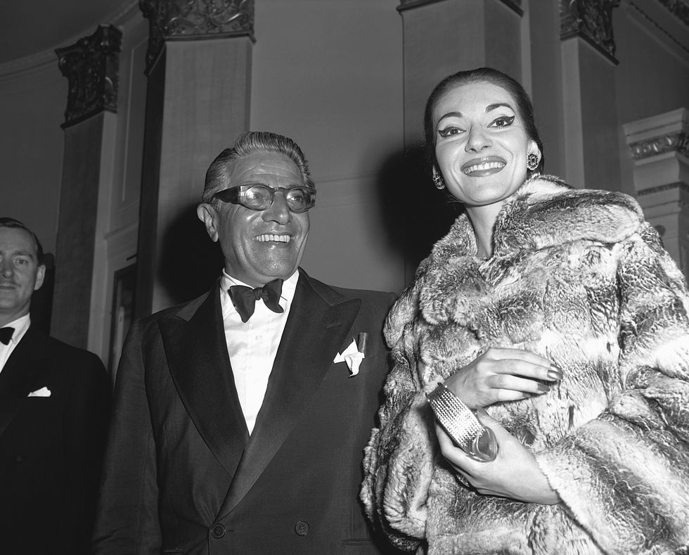 aristotle onassis and maria callas smile while standing next to each other, he wears a tuxedo with a bow tie and pocket square with glasses, she wears a large fur coat and holds a purse