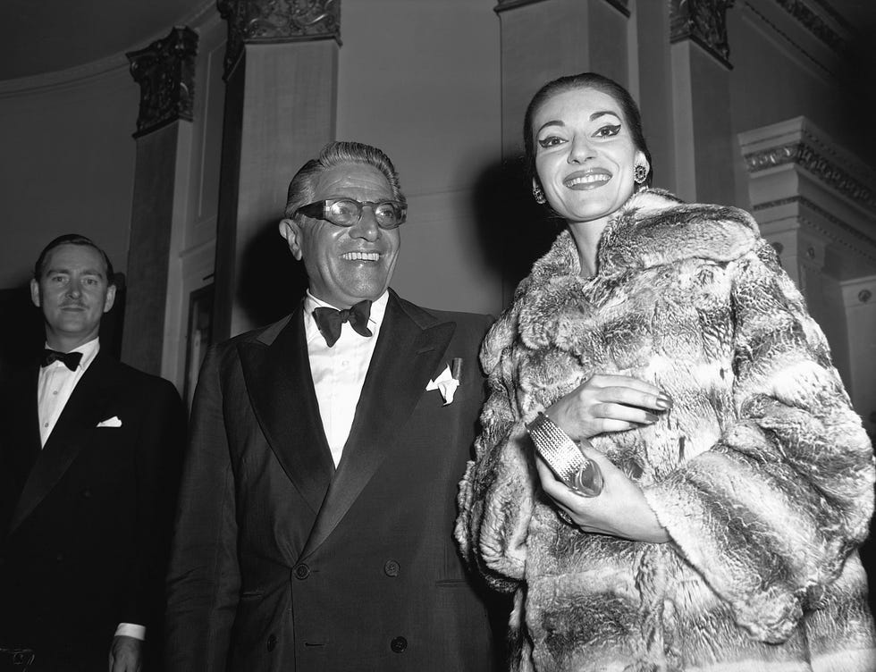 aristotle onassis and maria callas smile while standing next to each other, he wears a tuxedo with a bow tie and pocket square with glasses, she wears a large fur coat and holds a purse