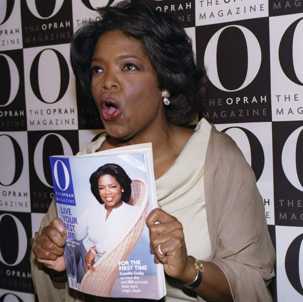 oprah winfrey holds the first issue of o, the oprah magazine while posing in front of a backdrop with the magazine's logo printed in black and white, she is wearing a cream colored dress, taupe shawl, jewelry and makeup