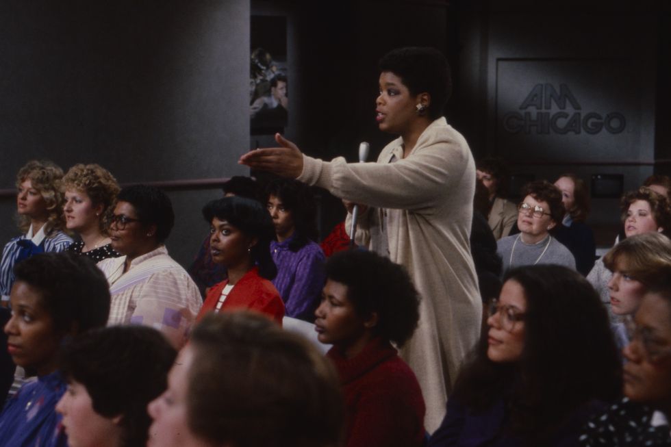 oprah winfrey stands in the middle of a seated tv audience while holding a microphone in one hand and extending her arm and pointing her hand toward the left of the frame, in the background is a logo for am chicago on the gray wall