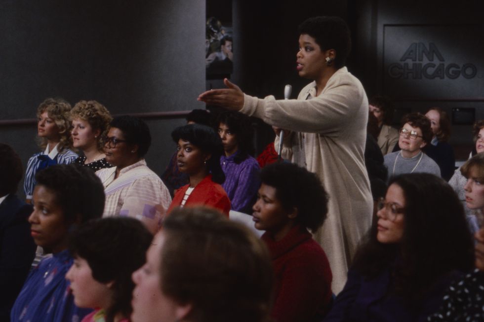 oprah winfrey stands in the middle of a seated tv audience while holding a microphone in one hand and extending her arm and pointing her hand toward the left of the frame, in the background is a logo for am chicago on the gray wall