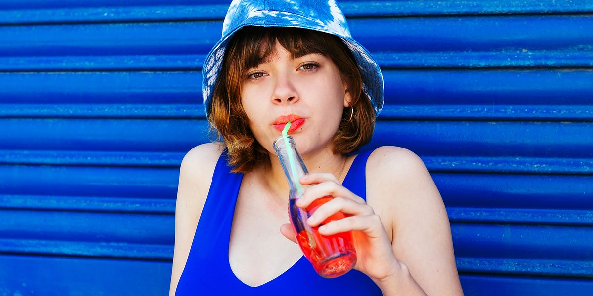 girl in bucket hat wearing orange lipstick and sipping on soda