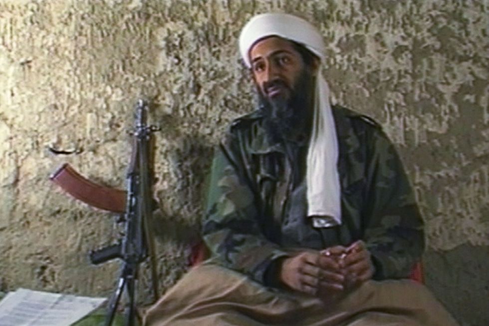osama bin laden sits against a concrete wall and appears to be talking, he wears a white head covering, green camo jacket, and camel colored blanket on his lap, to the left rest papers and a machine gun