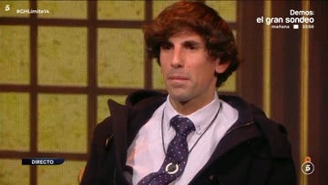 individual seated in front of a patterned backdrop wearing a tie and jacket