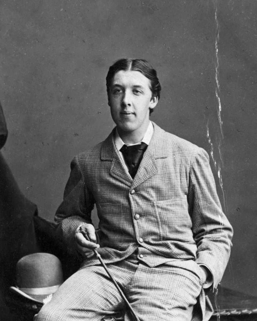 seated portrait of oscar wilde, he wears a plaid suit and holds a cane in one hand