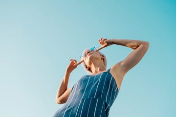 woman in a blue jumpsuit striking a playful pose, pointing at her stylish glasses, under a bright blue sky, radiating fun and confidence
