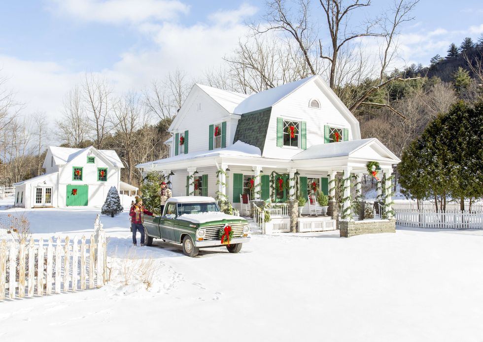 An old white farmhouse with a wreath wrapped around the porch pillars, a wreath on the window, and a vintage truck with a wreath on the bumper parked in the snow