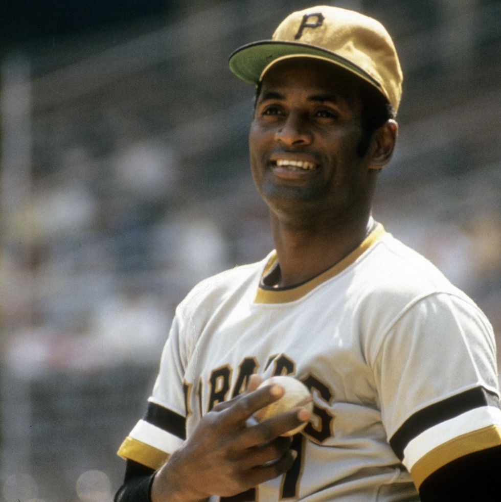 roberto clemente smiles and looks off into the distance, he wears a pittsburgh pirates uniform and yellow baseball cap and holds a baseball to his chest