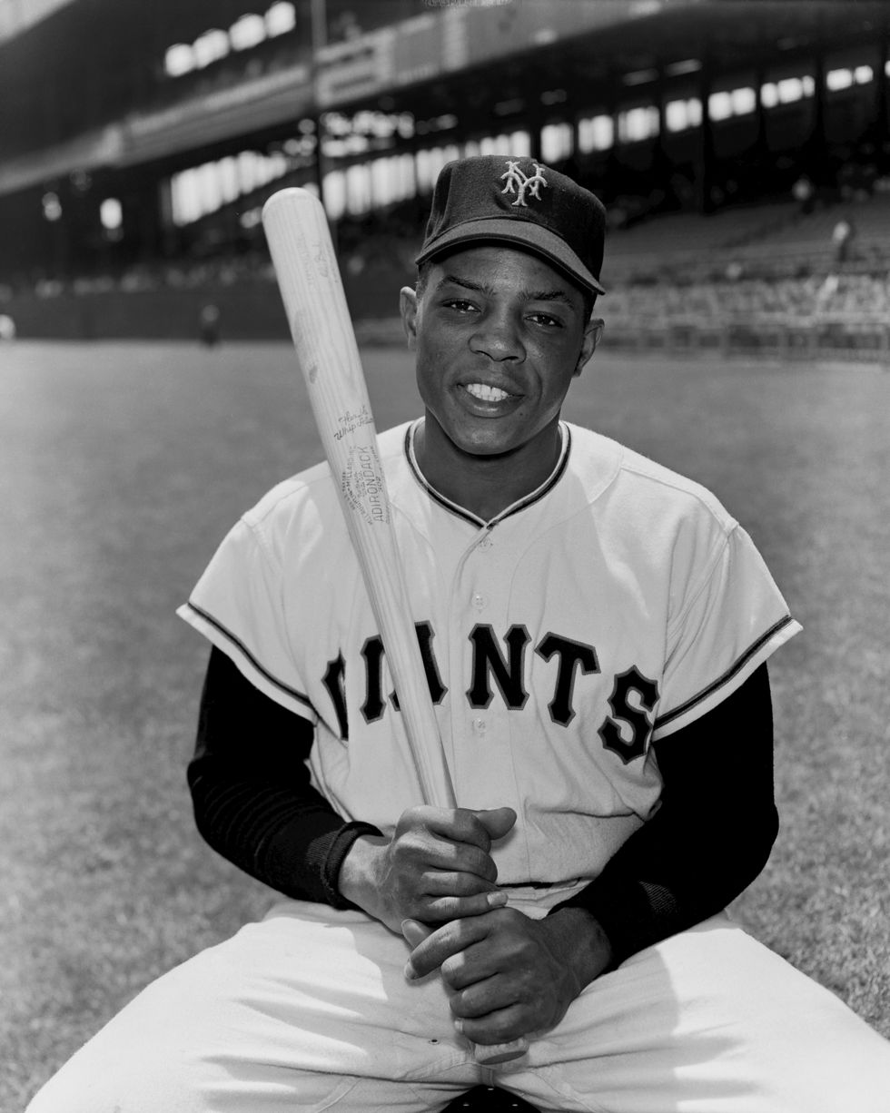 willie mays smiles at the camera as he sits inside a baseball stadium on the field, he holds a baseball bat with both hands and wears a uniform and baseball hat for the new york giants