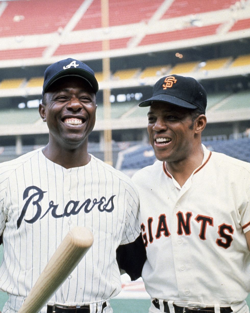 hank aaron and willie mays stand on a baseball field and smile, the men wear baseball uniforms and hats, aaron holds a bat in front of him