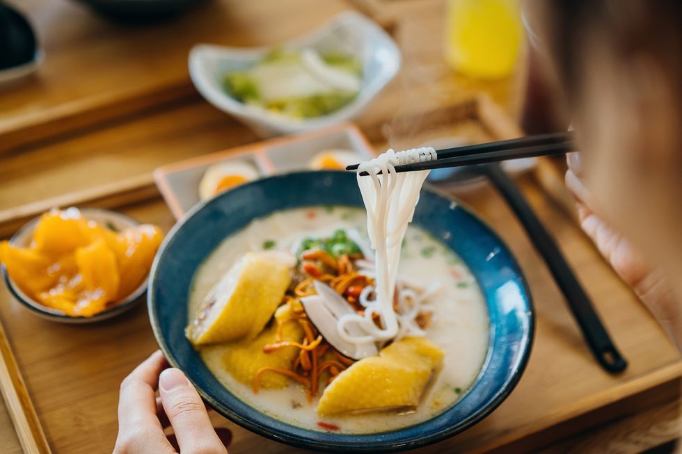 over the shoulder view of asian woman eating a bowl of freshly made noodle soup with chicken and vegetables with chopsticks in restaurant, served on a wooden tray asian food culture
